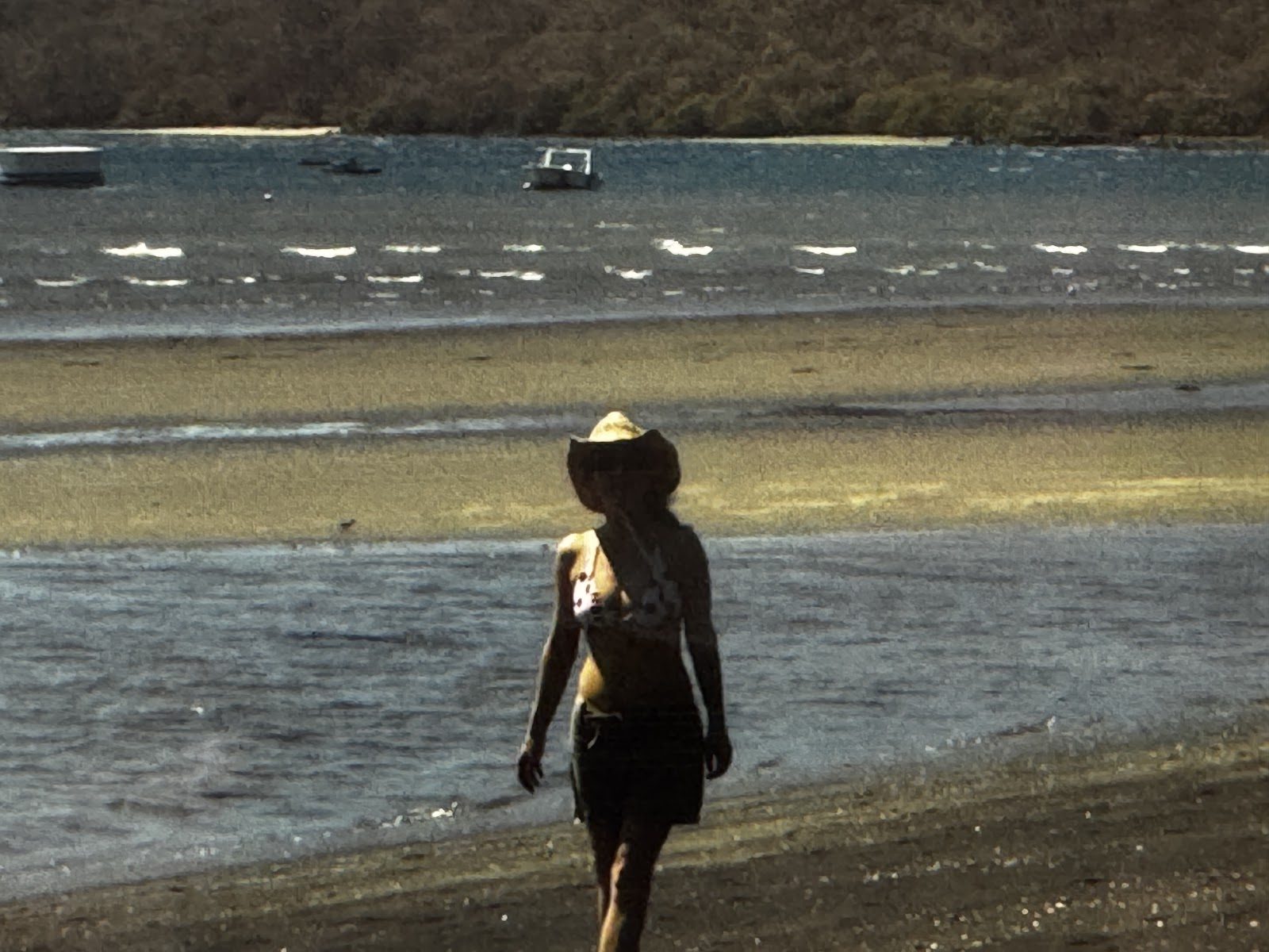 Laura walking alone along a shoreline in a sun hat
